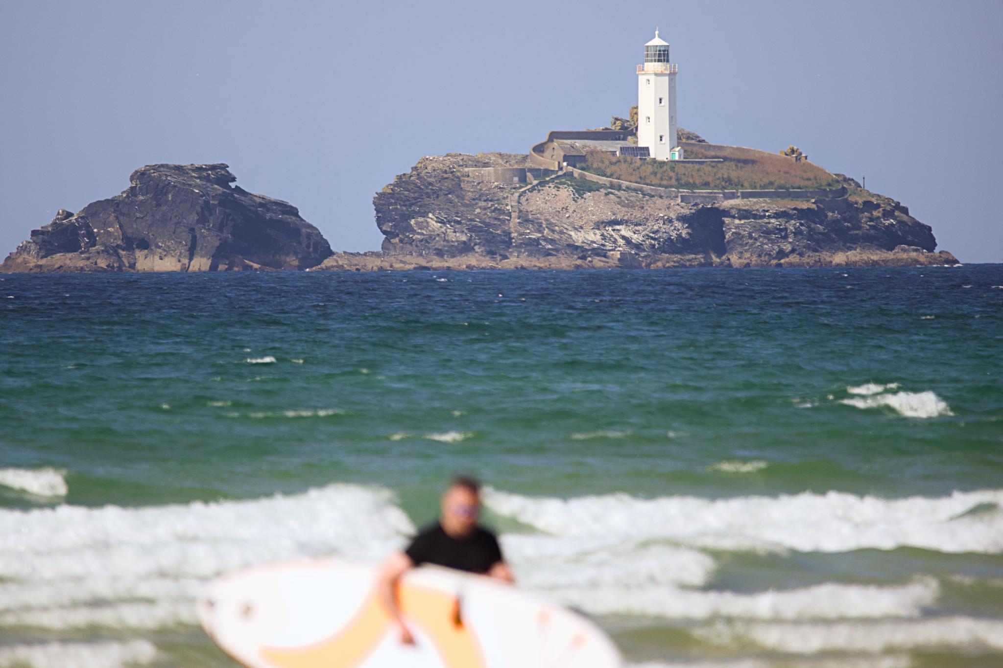 Godrevy lighthouse on its island, a surfer carrying a board in the foreground