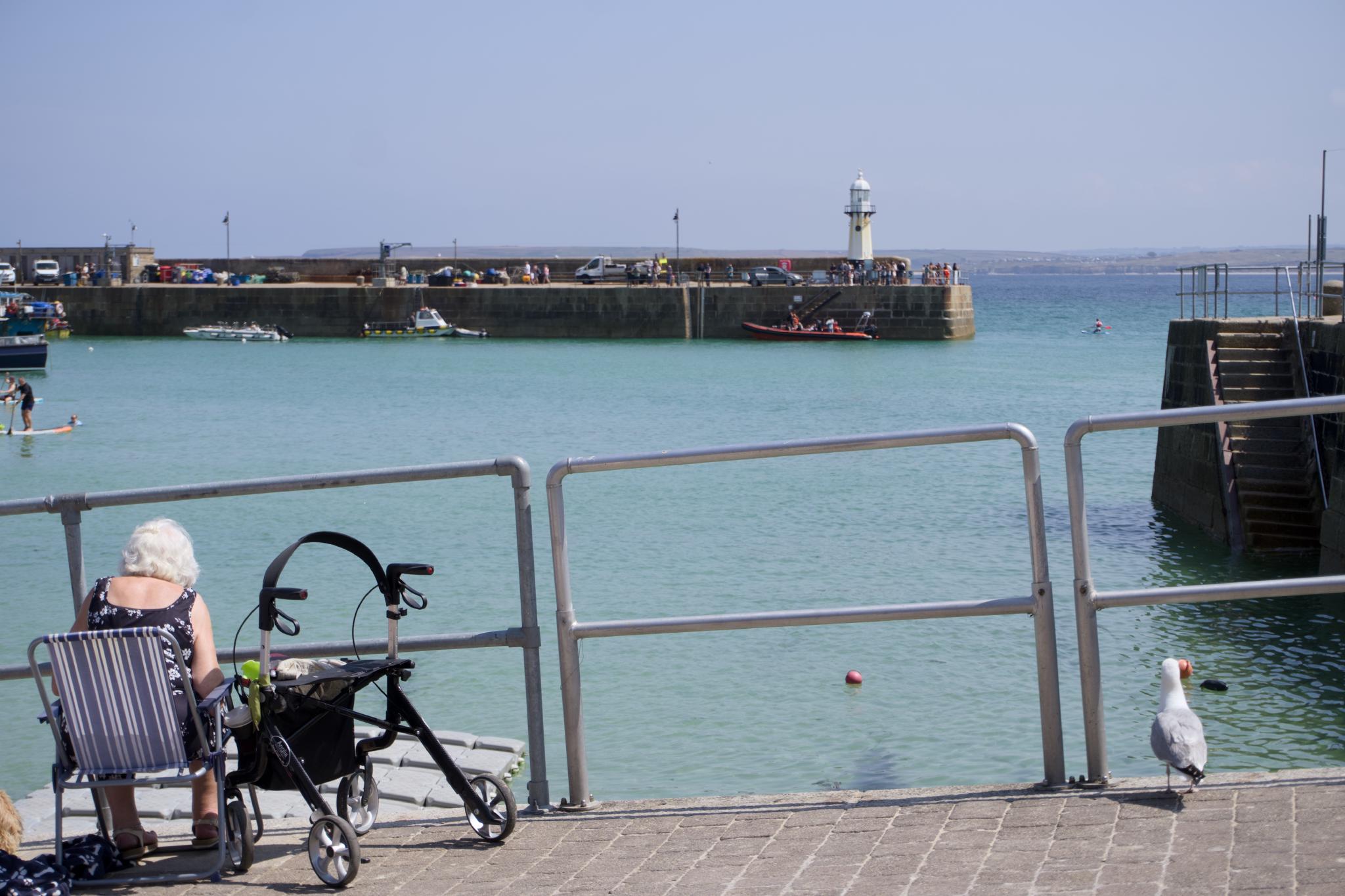 Woman with a rollator sitting at the harbour wall, the pier and lighthouse behind