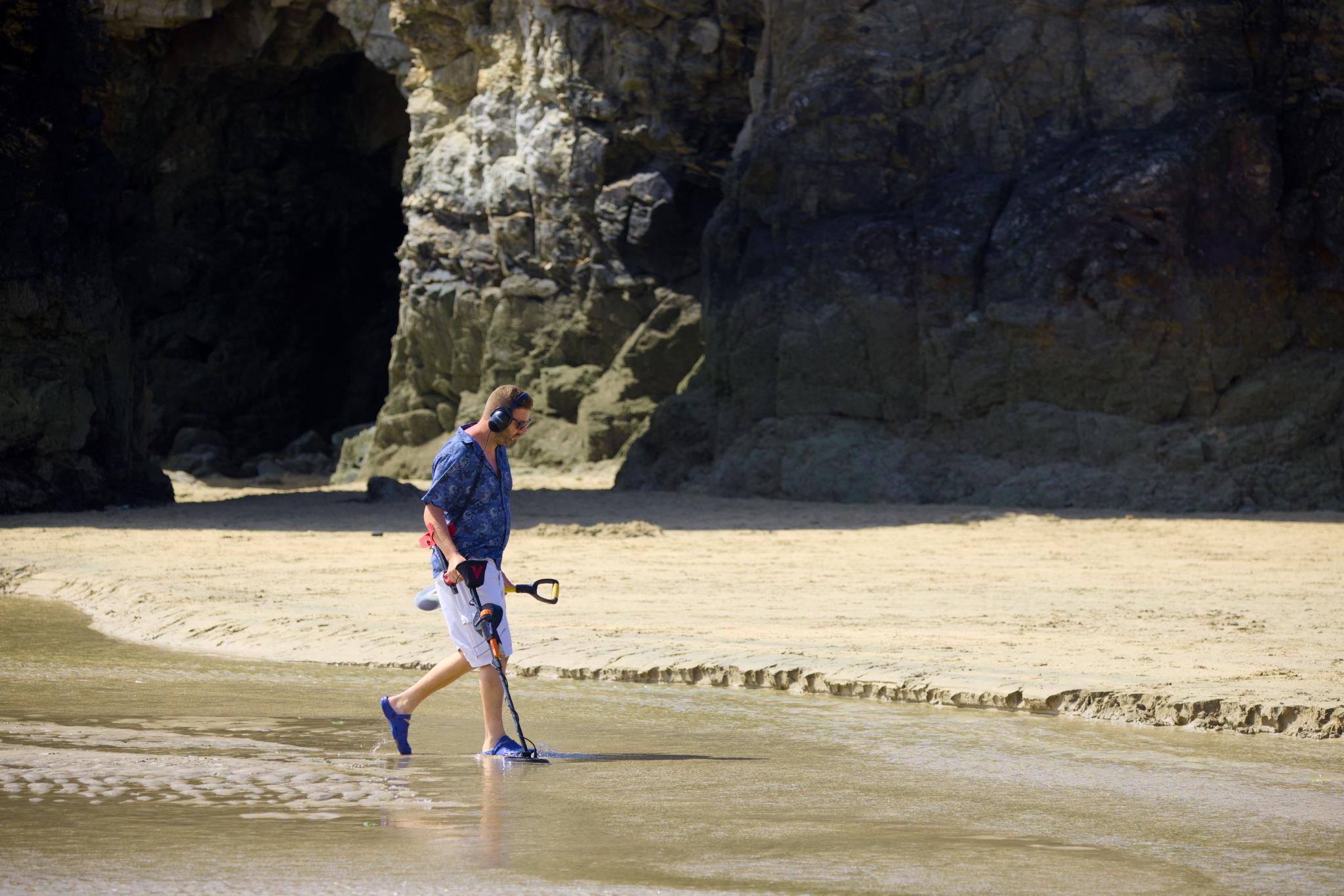 Man sweeping the wet sand with a metal detector in front of the cliffs