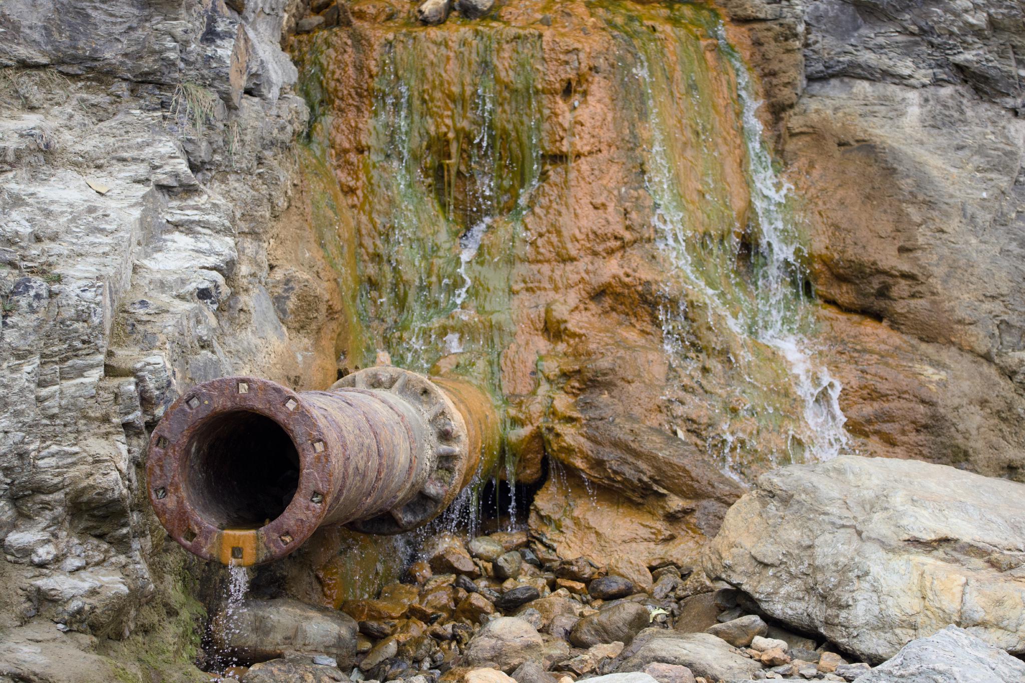 Rusty pipe below an orange-stained cliff face with water running down