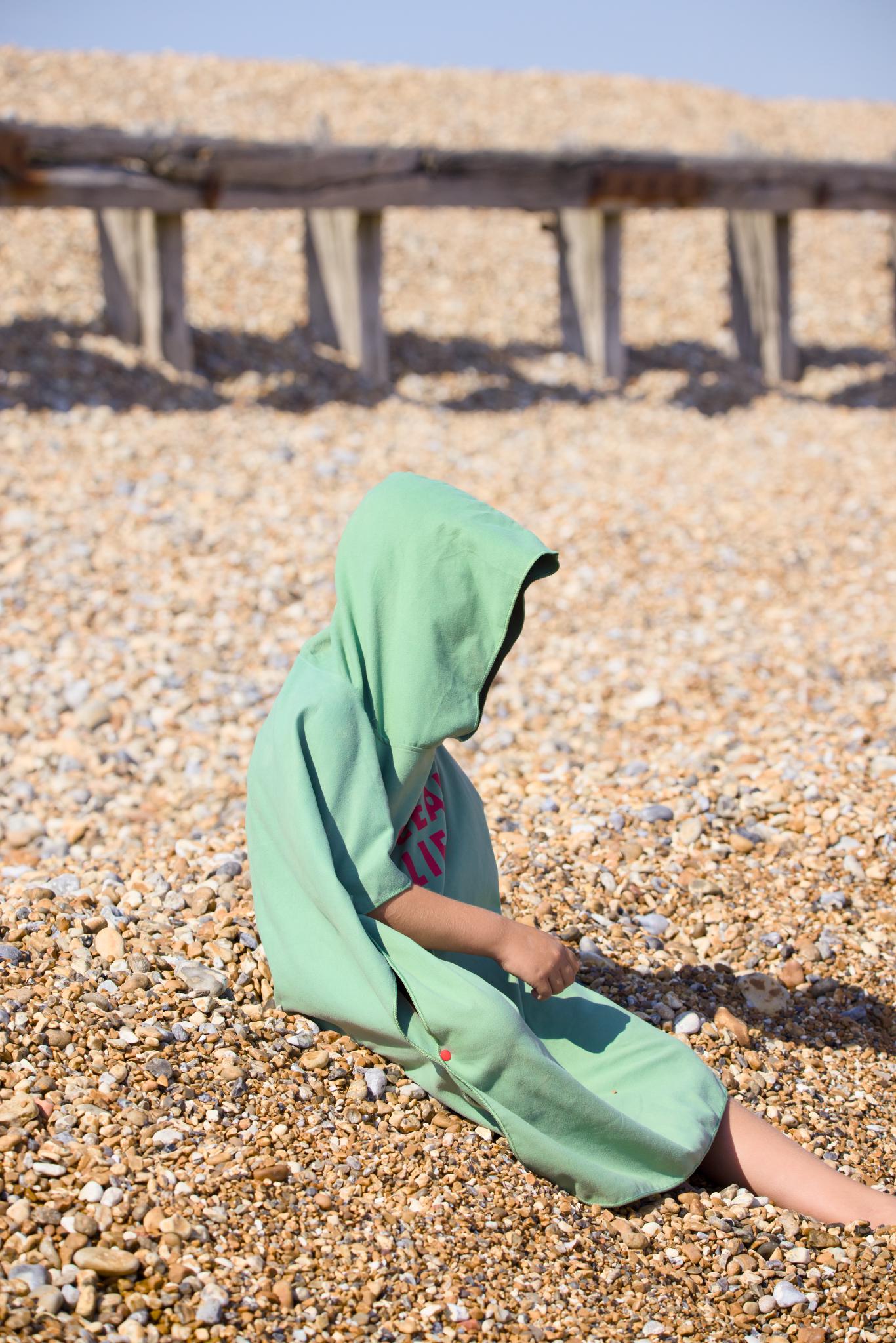 Boy in a green hooded towel poncho sitting on the shingle beach