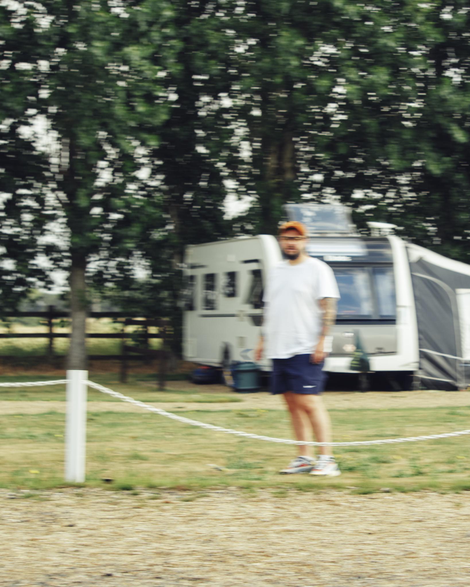 Sven at the rope fence of the pitch, slightly out of focus, a caravan behind