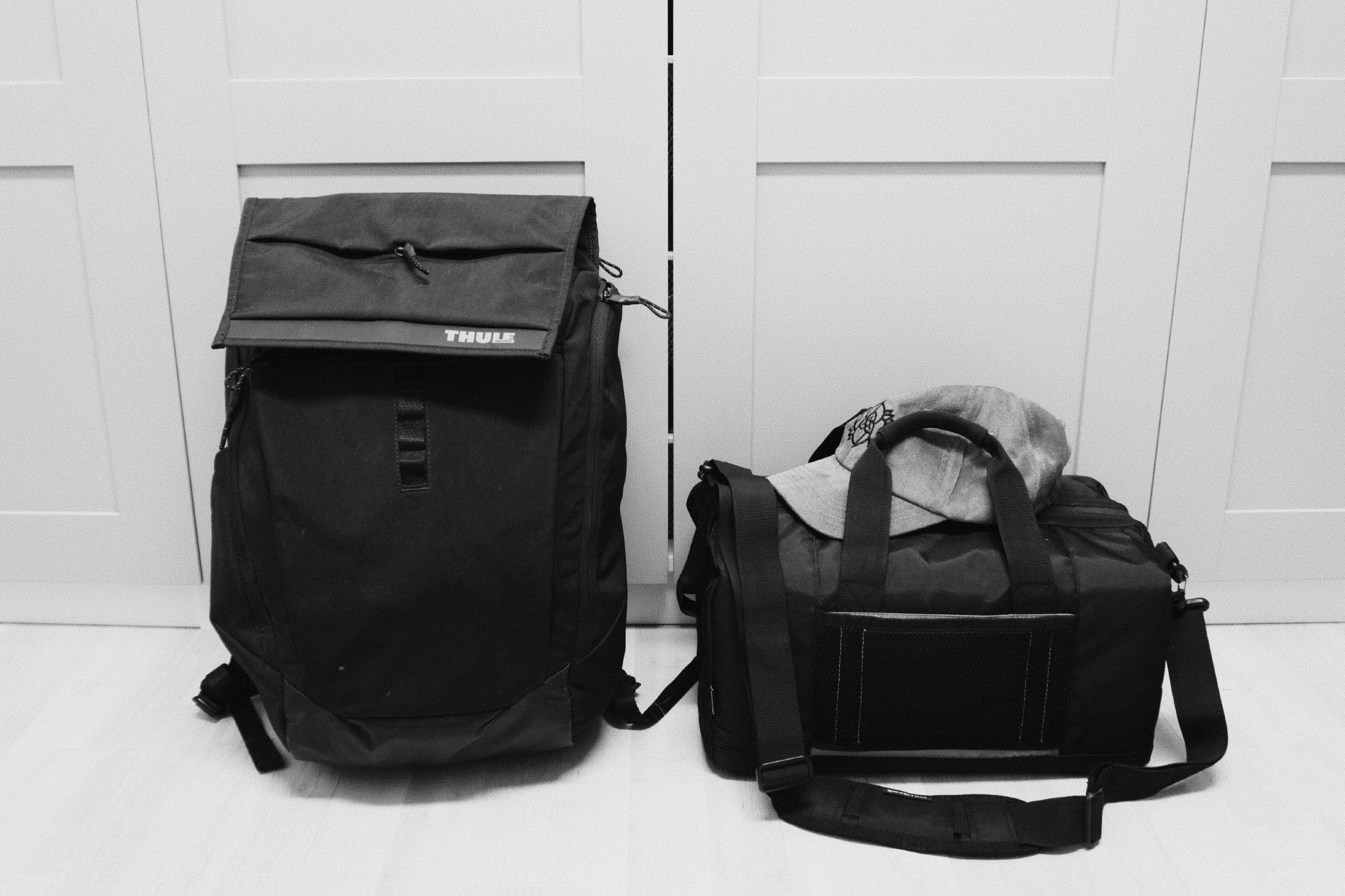 Black and white photo of a black Thule backpack and a black duffel bag with a grey cap on top, packed and standing side by side in front of white closet doors