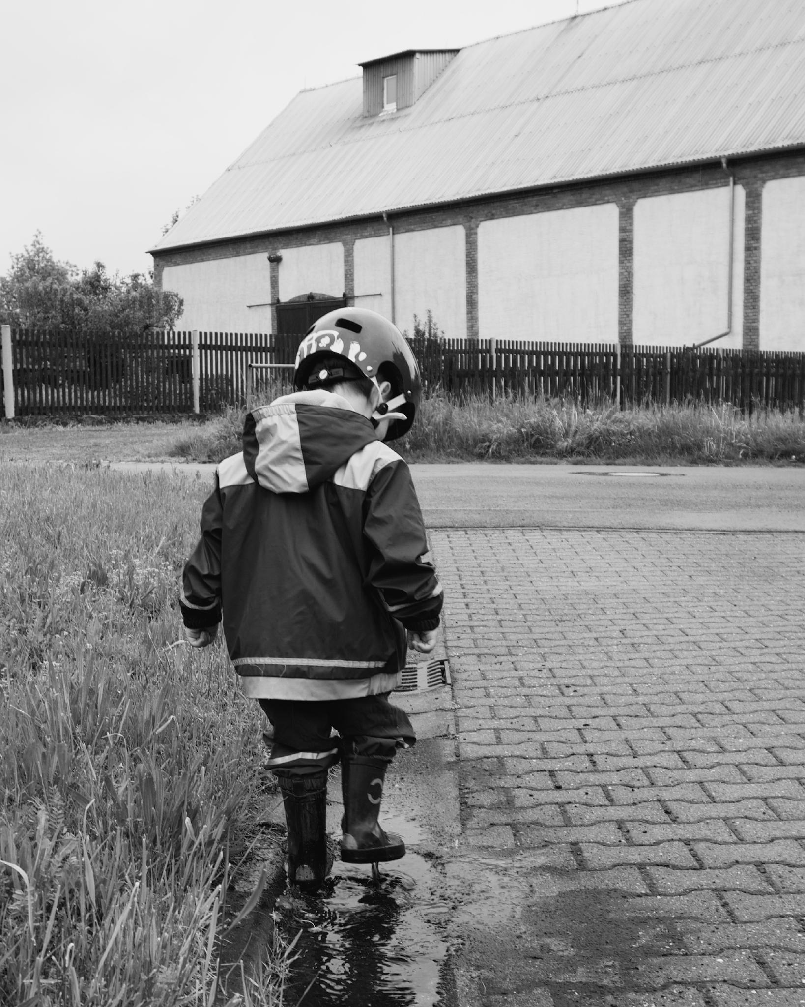 Black and white photograph of a small child in a raincoat, BONK helmet and rubber boots walking away through a puddle on a cobblestone path, a long barn with corrugated roof in the background