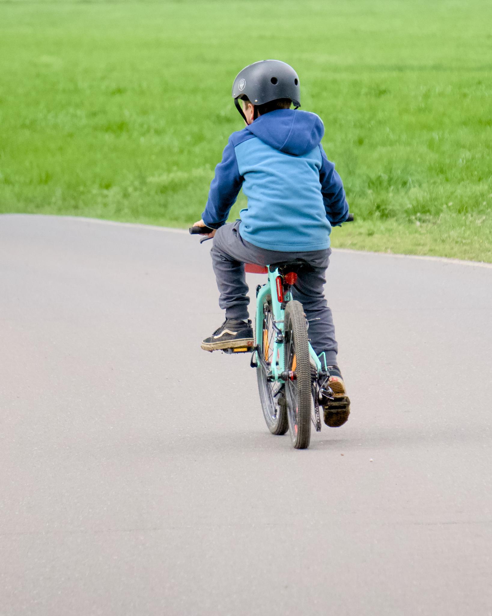 Young boy in a blue hoodie and black helmet riding a teal bicycle away from the camera on a paved path, with a grassy field in the background