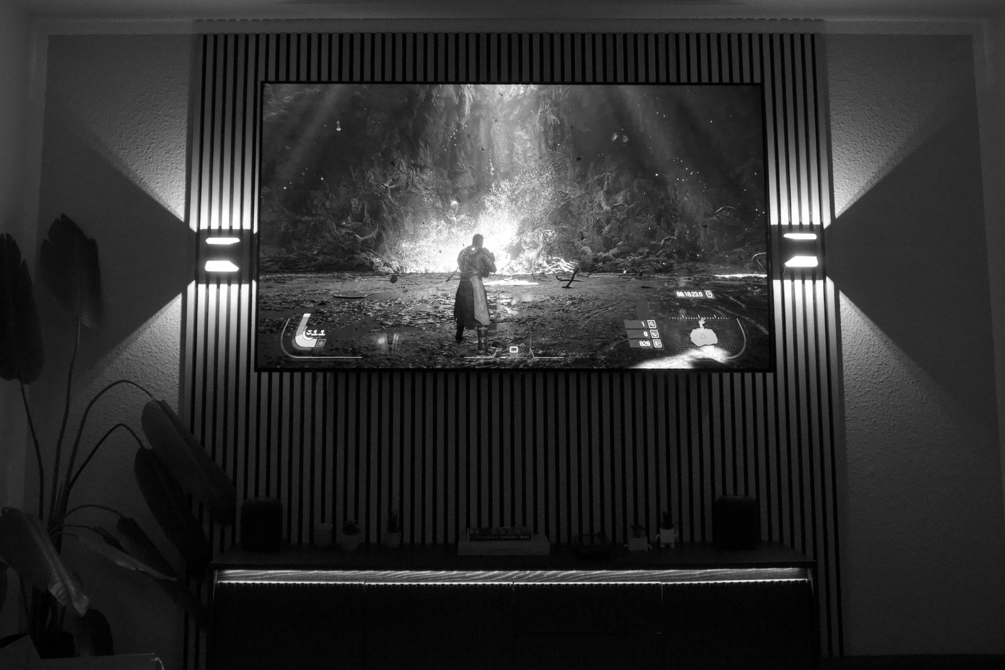 Black and white photograph of a wall-mounted TV showing the video game Saros, a cinematic scene with the protagonist standing in front of a glowing portal in a rocky landscape, the screen flanked by vertical wood slats and uplighters