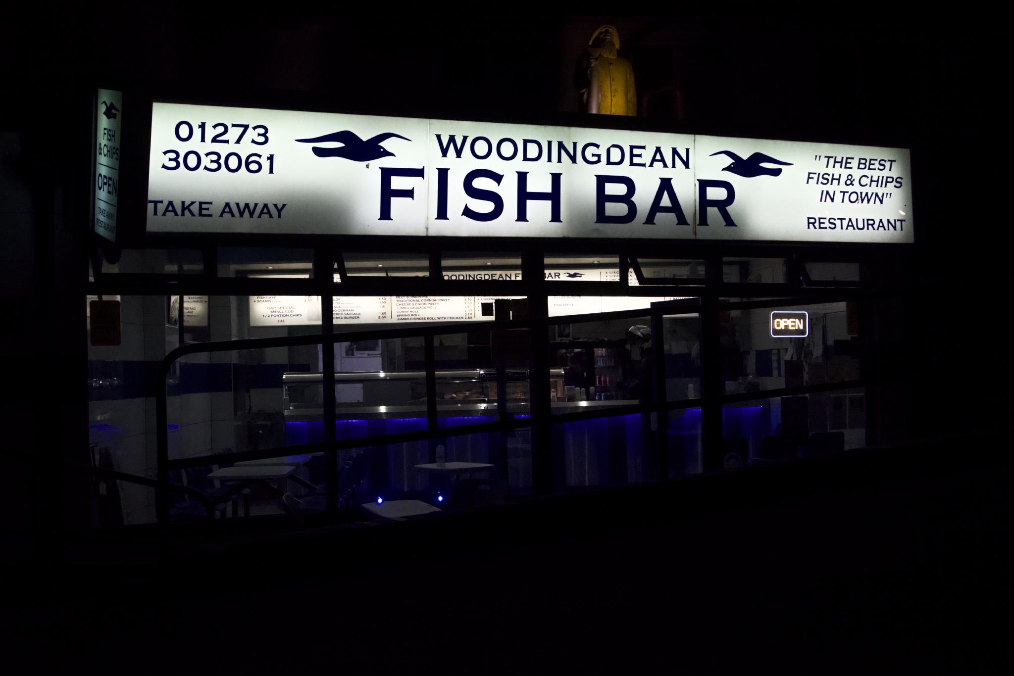 The illuminated sign of Woodingdean Fish Bar glowing at night, with two black seagull logos flanking the name and a dark empty interior visible through the windows